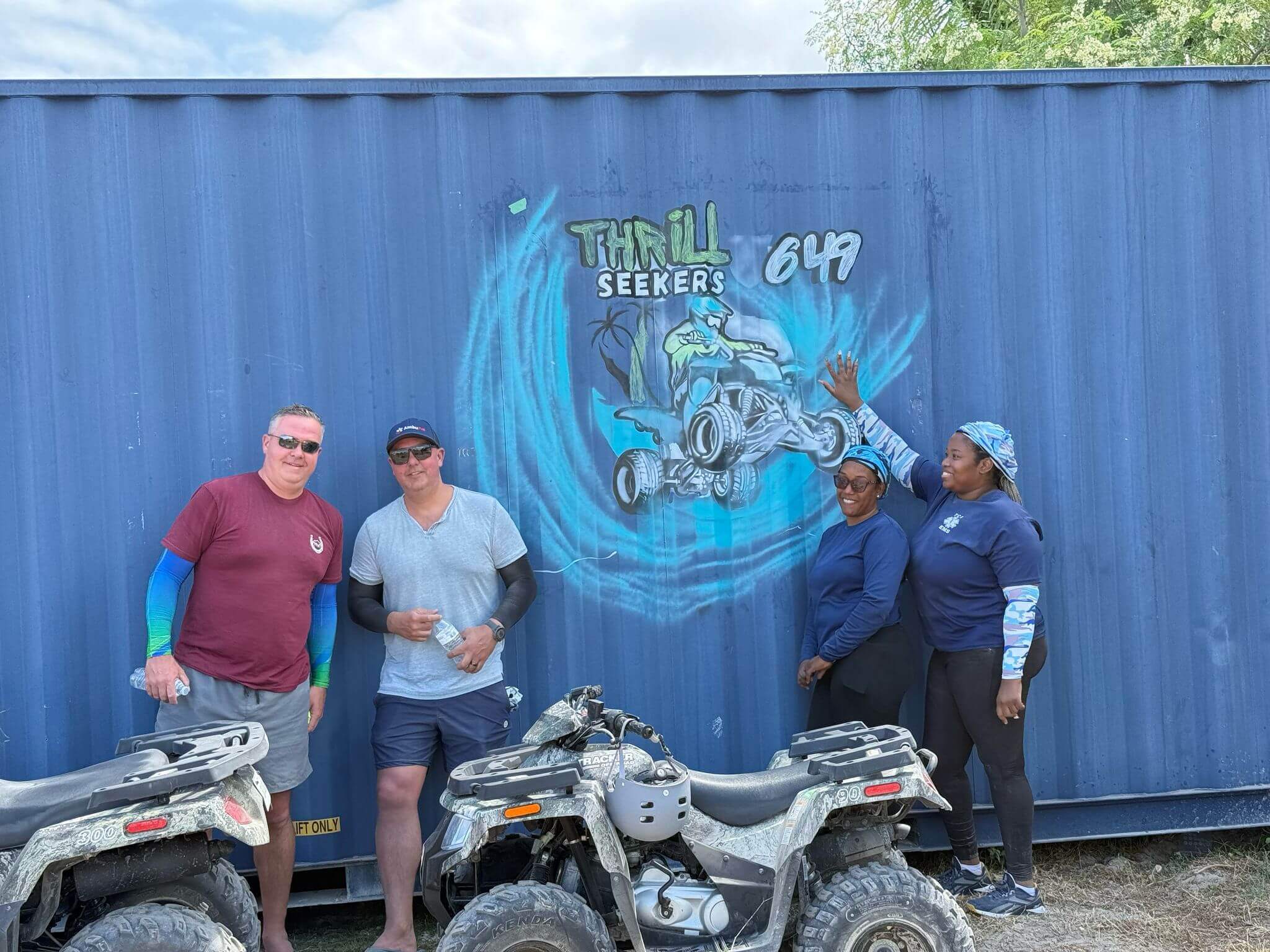 Group posing with ATVs in front of a blue container with "Thrill Seekers" graffiti, outdoor adventure theme.